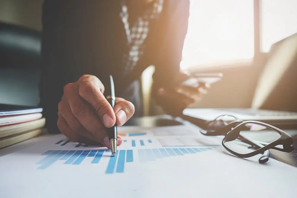 Person analyzing a bar chart with a pen, holding a phone, with glasses and documents on the table - accounting and payroll