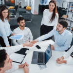 Team of professionals in a modern office shaking hands during a collaborative meeting, discussing reports and financial data, representing outsourced business valuation services and successful partnership.