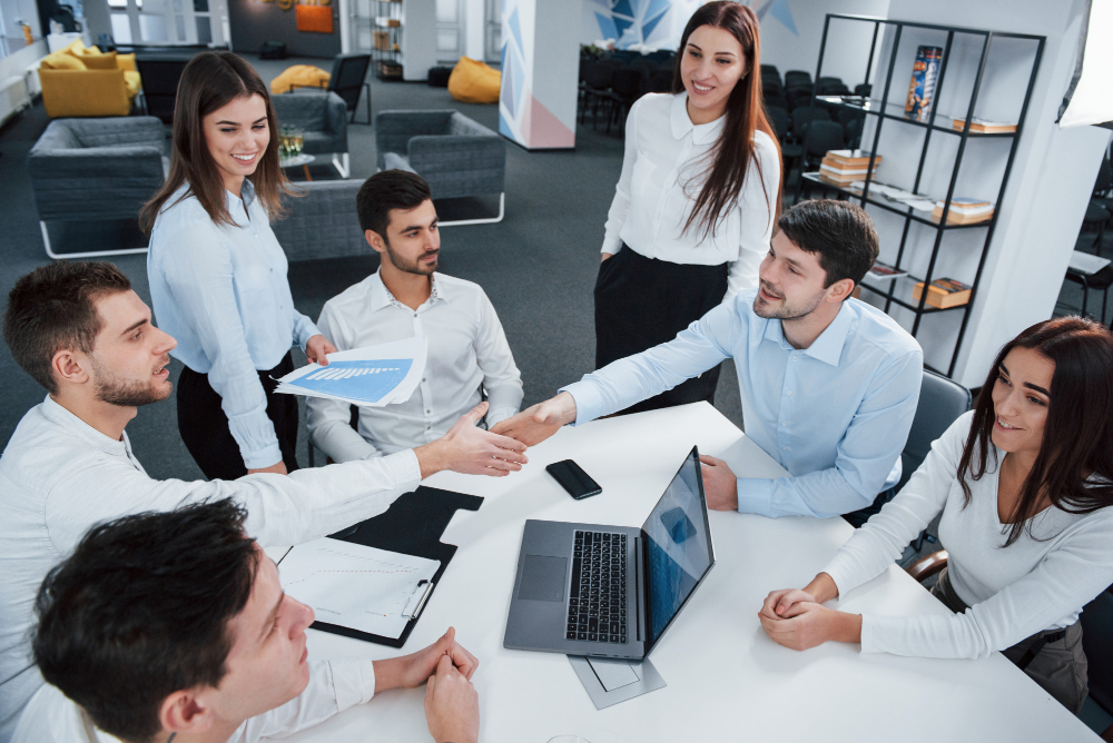 Team of professionals in a modern office shaking hands during a collaborative meeting, discussing reports and financial data, representing outsourced business valuation services and successful partnership.