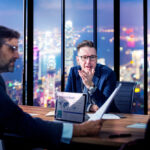 Business executives in a modern office reviewing reports and discussing company performance with a city skyline backdrop, highlighting strategic collaboration through Due Diligence Outsourcing.
