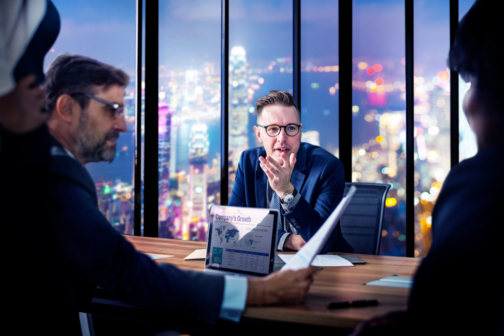Business executives in a modern office reviewing reports and discussing company performance with a city skyline backdrop, highlighting strategic collaboration through Due Diligence Outsourcing.